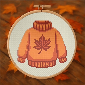 May include: Cross-stitch embroidery of an orange sweater with a maple leaf design. The sweater is in a wooden embroidery hoop. The background is a wooden surface with fall leaves.