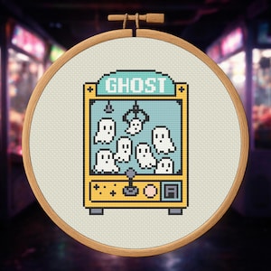 May include: A cross-stitch embroidery hoop featuring a pixelated arcade claw machine with the word "GHOST" above. The machine is yellow and blue, filled with white ghost figures. The embroidery is set against a neutral fabric.