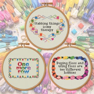 Puede incluir: Tres aros de punto de cruz con citas humorísticas. Uno dice "Stabbing things is my therapy", otro "One more row" y el último "Buying floss and using floss are two different hobbies". Los aros están rodeados de hilo de bordar colorido.