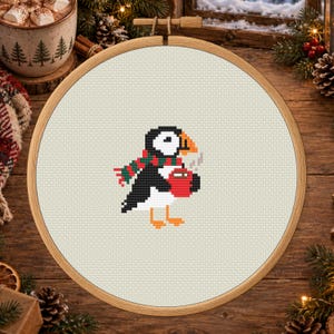 May include: A circular wooden embroidery hoop containing a cross-stitch pattern of a puffin. The puffin is black, white, and orange, wearing a red and green striped scarf and holding a red mug with steam rising from it, centered on cream fabric.