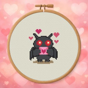 May include: Cross-stitch embroidery in a wooden hoop. The design features a black bat with red eyes and a pink heart-shaped letter. Pink hearts float around the bat. The background is a light beige fabric.