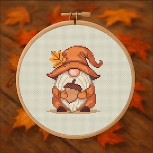 May include: A cross-stitch embroidery of a gnome holding an acorn, set in a wooden hoop. The gnome wears an orange hat and holds the acorn. The background is a light beige fabric, with a wooden frame and autumn leaves.