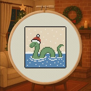May include: Cross-stitch embroidery in a wooden hoop. The design features a green Loch Ness Monster wearing a Santa hat, emerging from blue water with white snowflakes. The background is a light beige.