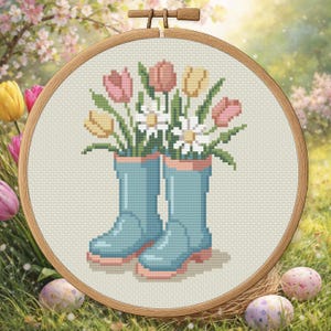 May include: Cross-stitch embroidery featuring blue wellington boots filled with pink, yellow, and white flowers. The design is set in a wooden hoop, with a spring background of flowers and Easter eggs.