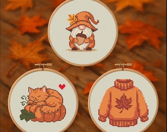 May include: Three cross-stitch embroidery hoops with autumn-themed designs. One features a gnome holding an acorn, another a sleeping cat on a pumpkin, and the last a sweater with a maple leaf. The hoops are set against a background of fall leaves.