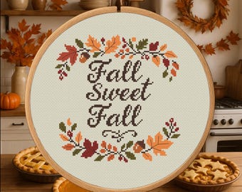 May include: A cross-stitch embroidery hoop with the words "Fall Sweet Fall" surrounded by a wreath of autumn leaves in orange, red, and green. Two pies are on the wooden table.