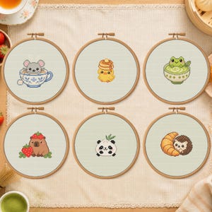 May include: Six circular wooden embroidery hoops containing cross-stitch patterns on light fabric. Designs include a mouse in a teacup, a chick with pancakes, a frog in a bowl, a capybara with strawberries, a panda dumpling, and a hedgehog with a croissant.
