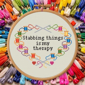 May include: A cross-stitch embroidery hoop with the text "Stabbing things is my therapy" surrounded by colorful thread spools and needles. The hoop is set against a backdrop of various colored embroidery floss.