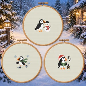 Puede incluir: Tres bastidores de bordado circulares de madera con diseños de punto de cruz sobre tela color crema. Uno muestra un frailecillo con un muñeco de nieve, otro un frailecillo patinando con copos de nieve y el tercero un frailecillo con gorro rojo.