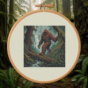 May include: A cross-stitch pattern in a round wooden embroidery hoop featuring a pixelated brown Bigfoot figure walking through a forest with tall trees, a fallen log, and a small waterfall in the background, rendered in shades of green and brown.
