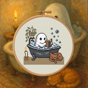 Puede incluir: Bordado de punto de cruz en un aro de madera que representa un fantasma relajándose en una bañera, leyendo un libro, con burbujas y un gato durmiendo. También se ven una vela encendida y una pequeña planta.