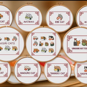 Peut inclure: Plusieurs cerceaux brodés avec des chats et des sushis. Chaque cercle a un fond blanc, un cadre rouge et un nom. Les cerceaux sont présentés sur une surface en bois, avec des baguettes visibles.