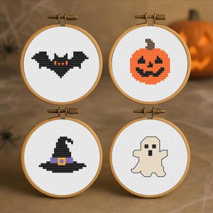 Può includere: Quattro telai da ricamo circolari in legno contenenti tessuto bianco con disegni a punto croce. I motivi raffigurano un pipistrello nero, una zucca arancione, un cappello da strega nero con fascia viola e un fantasma bianco.