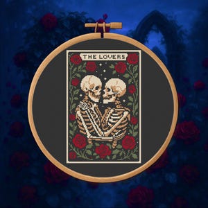 May include: A cross-stitch embroidery in a wooden hoop features a tarot card design. The card depicts two skeletons embracing, surrounded by red roses and green vines. The text "THE LOVERS" is at the top of the card.