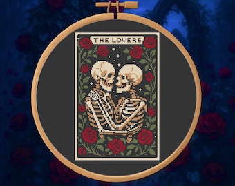 May include: A cross-stitch embroidery in a wooden hoop features a tarot card design. The card depicts two skeletons embracing, surrounded by red roses and green vines. The text "THE LOVERS" is at the top of the card.
