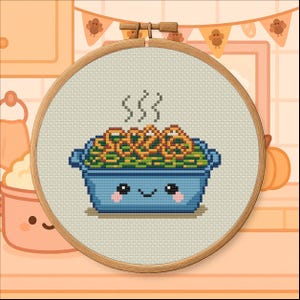 May include: Cross-stitch embroidery in a wooden hoop depicts a smiling blue casserole dish filled with green beans and fried onions, with steam rising. A Thanksgiving banner and pumpkin are in the background.