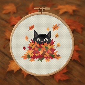 May include: Cross-stitch embroidery in a wooden hoop featuring a black cat peeking out from a pile of autumn leaves. The leaves are in shades of red, orange, and yellow, creating a fall theme. The background is a light beige fabric.