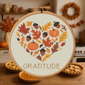 May include: A cross-stitch embroidery in a wooden hoop features a heart design filled with autumn leaves, pumpkins, and acorns in shades of orange, brown, and yellow. The word "GRATITUDE" is stitched below the heart. The background includes pies and fall decor.