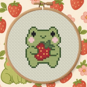 Könnte beinhalten: Kreuzstich-Stickerei in einem Holzrahmen mit einem grünen Frosch, der eine rote Erdbeere hält. Der Frosch hat rosa Wangen und schwarze Augen. Der Hintergrund ist hellbeige mit Erdbeer- und Blumenmustern.