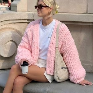 Soft plush puffy kawaii jacket in baby pink, chunky knit cardigan perfect for spring layering, woman sitting on a stone bench in a park wearing a cozy oversized hand-knit cardigan with soft plush texture.