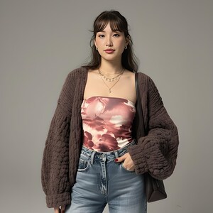 以下が含まれることがあります： 茶色の分厚いニットカーディガン、柄物のチューブトップ、青いジーンズを着た女性。チューブトップはピンクと白の雲のようなデザインです。彼女はまた、重ね着のネックレスとイヤリングを身に着けています。