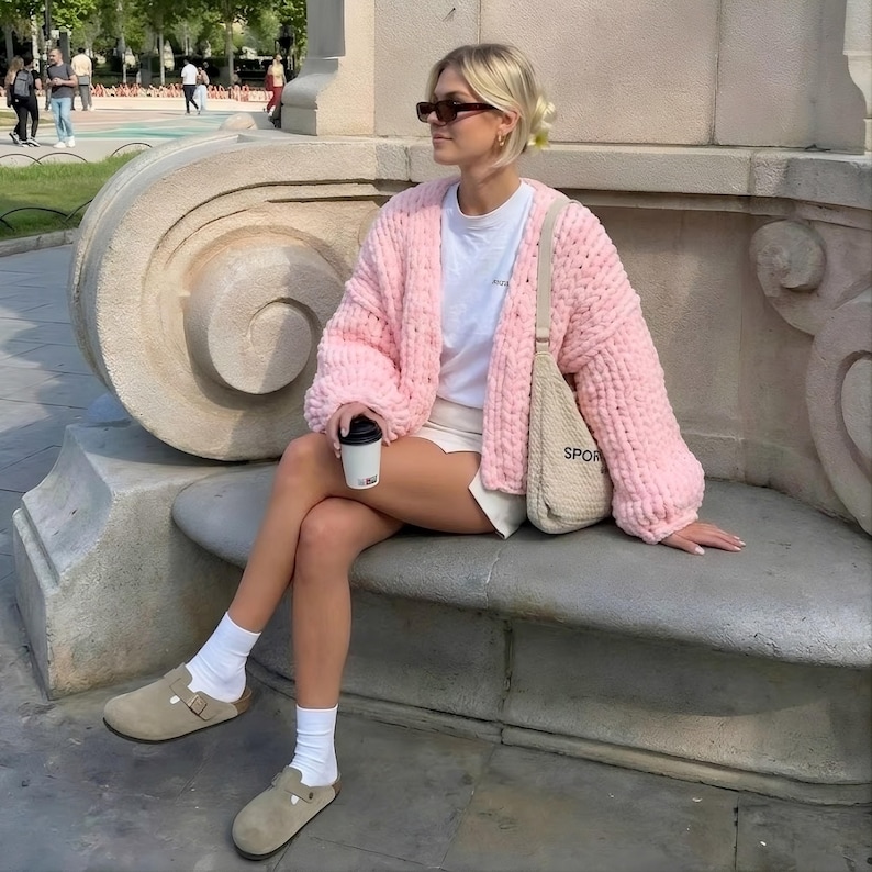 Soft plush puffy kawaii jacket in baby pink, chunky knit cardigan perfect for spring layering, woman sitting on a stone bench in a park wearing a cozy oversized hand-knit cardigan with soft plush texture.