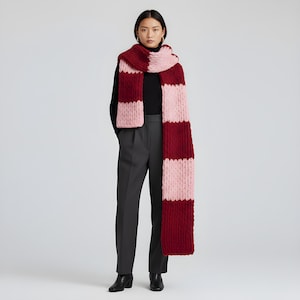 May include: A chunky knit scarf in a bold color block design. Alternating stripes of deep red and light pink create a striking visual. The scarf is thick and appears warm, suitable for cold weather. The model is wearing a black top and gray pants.
