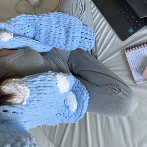 May include: A light blue, chunky knit sweater with white cloud-shaped accents. The sweater is made of a soft, textured yarn. The sleeves are visible, and the overall style is cozy and comfortable.
