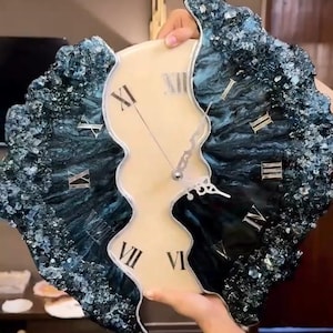 Puede incluir: Un reloj de pared único con una esfera color crema y números romanos. Los bordes exteriores del reloj son de color verde azulado oscuro con una apariencia texturizada y cristalina. El reloj es sostenido por una persona.
