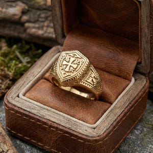 Peut inclure: Une bague dorée avec une face en forme de bouclier ornée d'une croix. La bague est présentée dans un écrin en cuir marron. La bague présente des détails complexes et une finition polie. La bague est de style chevalière, avec un design classique et élégant.
