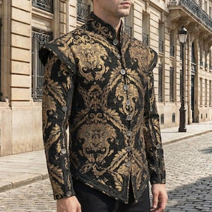 Op de afbeelding: Een zwart en goudkleurig brokaat jasje met een hoge kraag en sierlijke knopen. Het jasje heeft een gedetailleerd bloemmotief en gestructureerde schouders, wat een historische of theatrale uitstraling oproept.