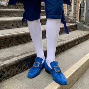 Op de afbeelding: Een paar felblauwe suède loafers met goudkleurige vierkante gespen. De schoenen worden gedragen met witte sokken en een donkerblauwe broek. De foto is buiten op een stenen trap gemaakt.