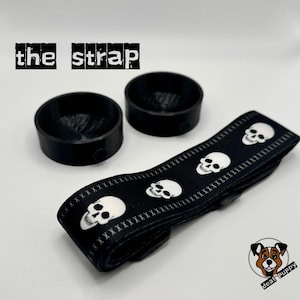 May include: Black and white skull patterned strap with the text "the strap" above it. Two black round objects are also in the image.