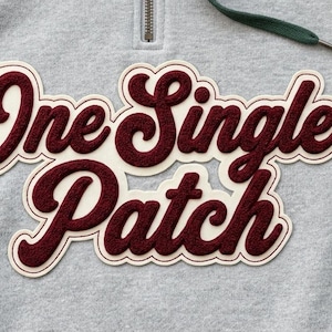Pode incluir: Close-up de um moletom cinza com um patch bordô e creme. O patch diz "One Single Patch" em uma fonte de script. O moletom tem um zíper e um cordão verde.