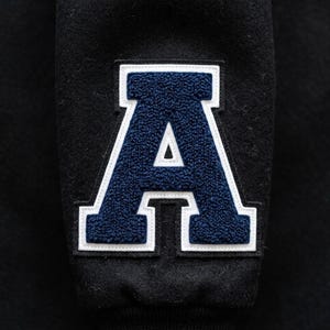 May include: A black wool-blend fabric sleeve featuring a large, centered letter A patch. The letter A is constructed with a textured, dark blue chenille center, outlined by a thick white border, and finished with a thin black stitched edge.
