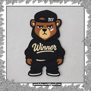 May include: A brown bear patch wearing a black hoodie with the word "Winner" in gold script. The bear is wearing a black cap with "NY" on it. The bear has a white mark under its eye and is wearing black pants and shoes.