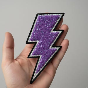 Może przedstawiać: Fioletowa błyskawica z brokatem z czarną obwódką i srebrnymi akcentami. Naszywka jest trzymana w dłoni na neutralnym tle. Tę ozdobną naszywkę można naprasować na ubrania lub akcesoria.