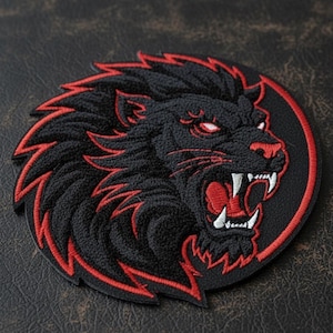 Puede incluir: Parche bordado con la cabeza de un león rugiendo. El león es negro con detalles rojos en la melena, la boca y los ojos, sobre un fondo circular negro con un borde rojo. El parche es redondo y tiene una apariencia texturizada.