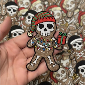 Könnte beinhalten: Ein gestickter Aufnäher in Form eines Lebkuchenmanns mit Totenkopf-Kopf, der eine rot-grün gestreifte Elfenmütze trägt. Der Körper zeigt weiße Knochenmuster, bunte Lichterketten um den Oberkörper und ein rotes Geschenk mit einer grünen Schleife.