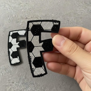 Puede incluir: Letra "F" en blanco y negro con diseño de balón de fútbol, adornada con brillantes pedrería. La letra es sostenida por una mano sobre un fondo gris. El diseño presenta patrones hexagonales en blanco y negro.