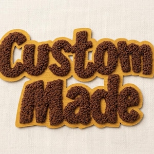 Könnte beinhalten: Ein Aufnäher mit den Worten Custom Made in dunkelbraunen, strukturierten, flauschigen Chenille-Buchstaben. Die Buchstaben sind auf einen senfgelben Filzhintergrund geschichtet, der der Kontur des Textes folgt und ein erhabenes Aussehen erzeugt.