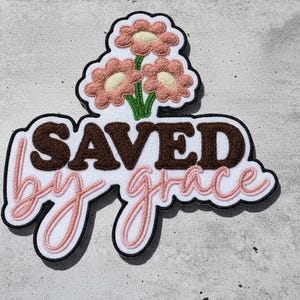 Puede incluir: Un parche decorativo con las palabras "SAVED by grace" en letras marrones y rosas. El parche presenta tres flores rosas con centros blancos y tallos verdes. El parche tiene un borde negro.