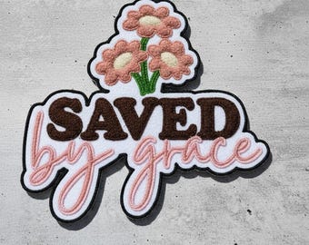 Op de afbeelding: Een decoratieve patch met de woorden "SAVED by grace" in bruine en roze letters. De patch heeft drie roze bloemen met witte centra en groene stelen. De patch heeft een zwarte rand.