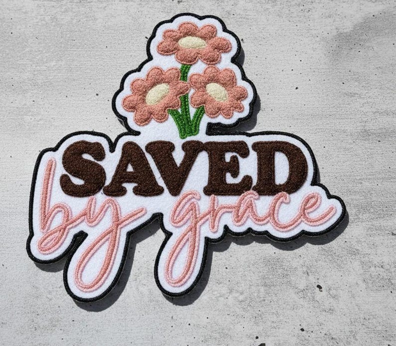 Pu&ograve; includere: Una toppa decorativa con le parole "SAVED by grace" in lettere marroni e rosa. La toppa presenta tre fiori rosa con centri bianchi e steli verdi. La toppa ha un bordo nero.