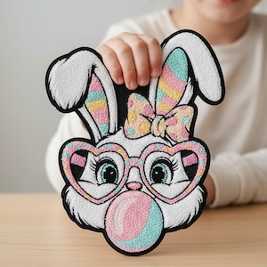 Puede incluir: Un parche de conejo blanco con contorno negro, con gafas en forma de corazón y soplando una burbuja de chicle. El conejo tiene un lazo y orejas a rayas de colores. El parche está sostenido por una persona.