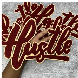 Op de afbeelding: Bordeauxrode en crèmekleurige viltpatches met het woord "Hustle" in een cursief lettertype. De patches worden in een hand gehouden, met het woord "Hustle" prominent weergegeven. De patches hebben een varsity letter stijl.