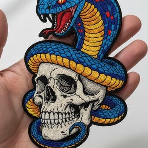 May include: Embroidered patch featuring a skull with a blue and yellow snake wrapped around it. The snake has red accents and is poised to strike. The patch is a detailed, colorful design.