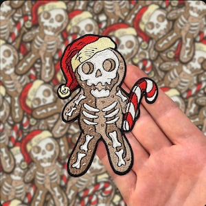 Könnte beinhalten: Ein gestickter Aufnäher, der einen Lebkuchenmann mit Skelett-Design zeigt, der eine rot-weiße Weihnachtsmannmütze trägt und eine rot-weiß gestreifte Zuckerstange hält. Der Aufnäher hat einen braunen Lebkuchenkörper mit weißen Knochendetails.