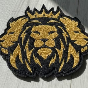 Puede incluir: Parche bordado con un diseño de león dorado y negro. El parche representa una cara de león central con una corona, flanqueada por dos caras de león adicionales. El diseño tiene una apariencia texturizada y en relieve.