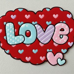 Puede incluir: Un parche rojo en forma de corazón con la palabra "LOVE" en letras de burbujas. Las letras son de color azul claro con lunares blancos, moradas con rayas diagonales y rosas con lunares blancos. Pequeños corazones rosas y azules están dispersos sobre el fondo rojo.